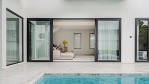 Un área moderna junto a la piscina con vistas directas a una sala de estar a través de amplios ventanales.