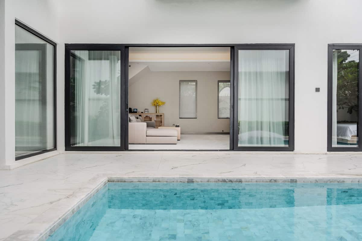 Un área moderna junto a la piscina con vistas directas a una sala de estar a través de amplios ventanales.