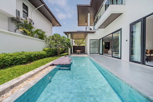 Zona de piscina residencial moderna amb una piscina de blau clar, una zona de seients coberta i vista a la casa.