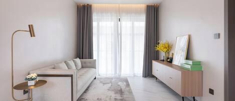 A modern living room with a sofa, a small side table, a floor lamp, a console table with decorative items, and a large window with curtains.