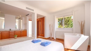 Ett modernt hotellrum med en stor säng, ett vitt badkar och ett stort fönster med utsikt över träd.