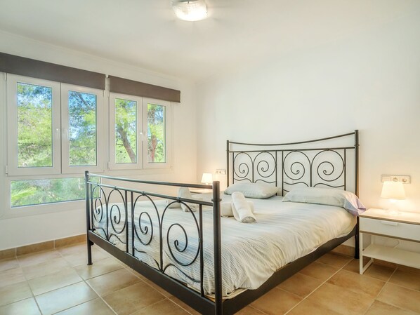 A bedroom with a metal bed frame, a nightstand, a lamp, and a window with a view of trees.
