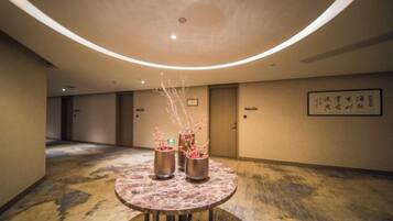 A hotel corridor with a round table featuring two metallic vases with branches, a curved ceiling with recessed lighting, and a framed sign on the wall.