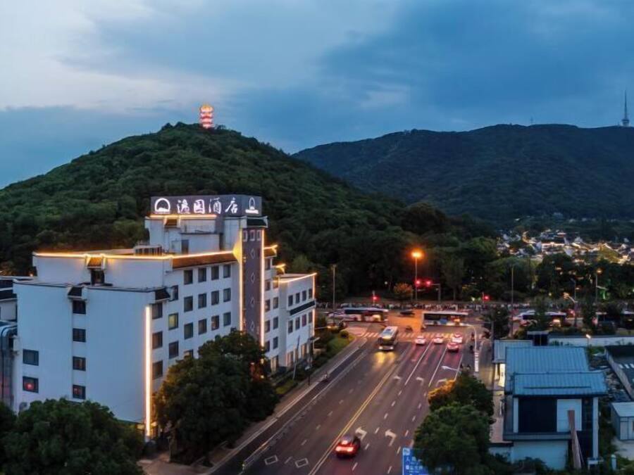 A multi-story building with a sign indicating a hotel. There is a road with vehicles in the foreground, and a hill with a tower in the background.