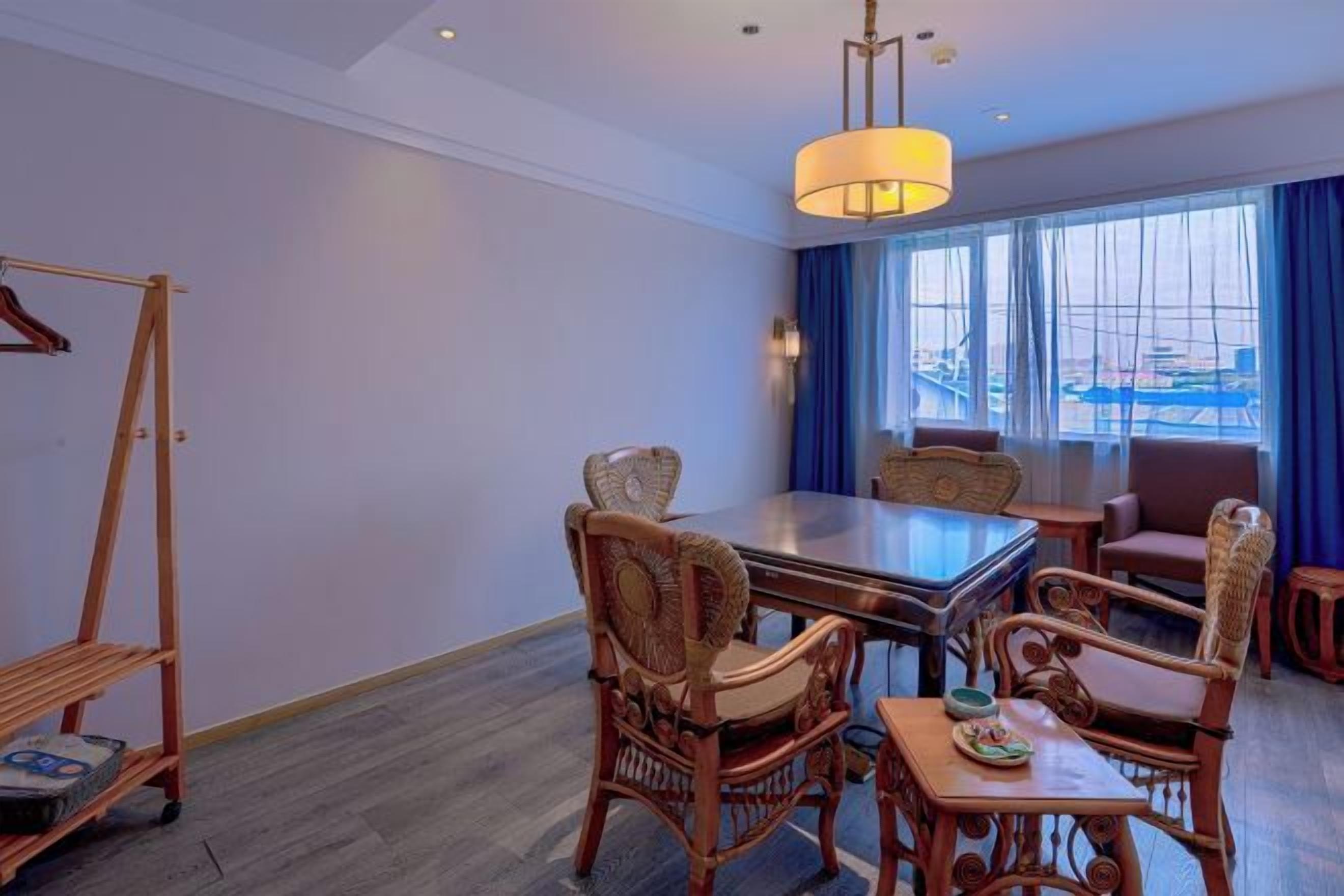 Habitación de hotel con mesa de comedor, sillas de mimbre, una pequeña mesita auxiliar, una lámpara colgante y vistas de la ciudad a través de amplias ventanas.