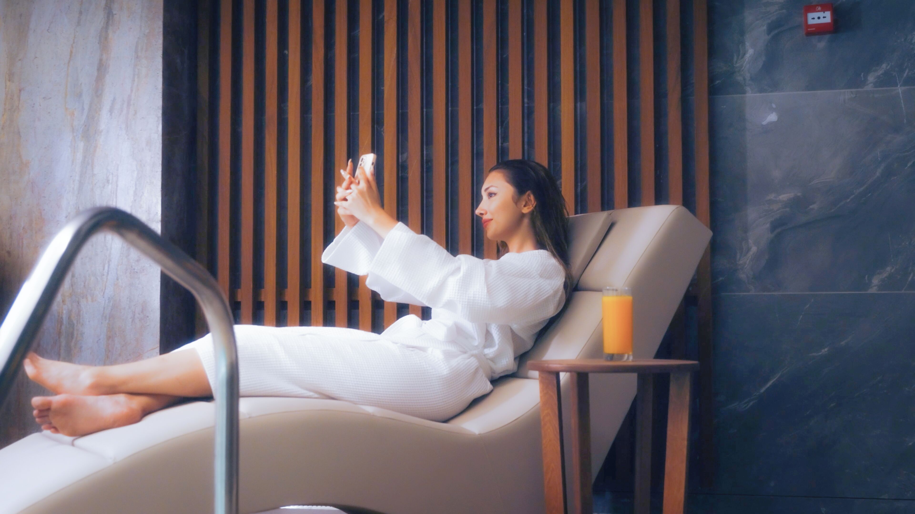 A person relaxing in a modern lounge chair with a smartphone, a glass of orange juice, and a wooden slat wall in the background.