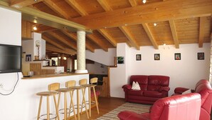 Sala de estar com sofá de couro vermelho, cadeiras de madeira, área de cozinha com balcão e teto de madeira.