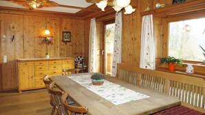 Uma sala de jantar de madeira com uma mesa, cadeiras e um banco. Há uma janela com cortinas, um radiador e um vaso com planta no peitoril.
