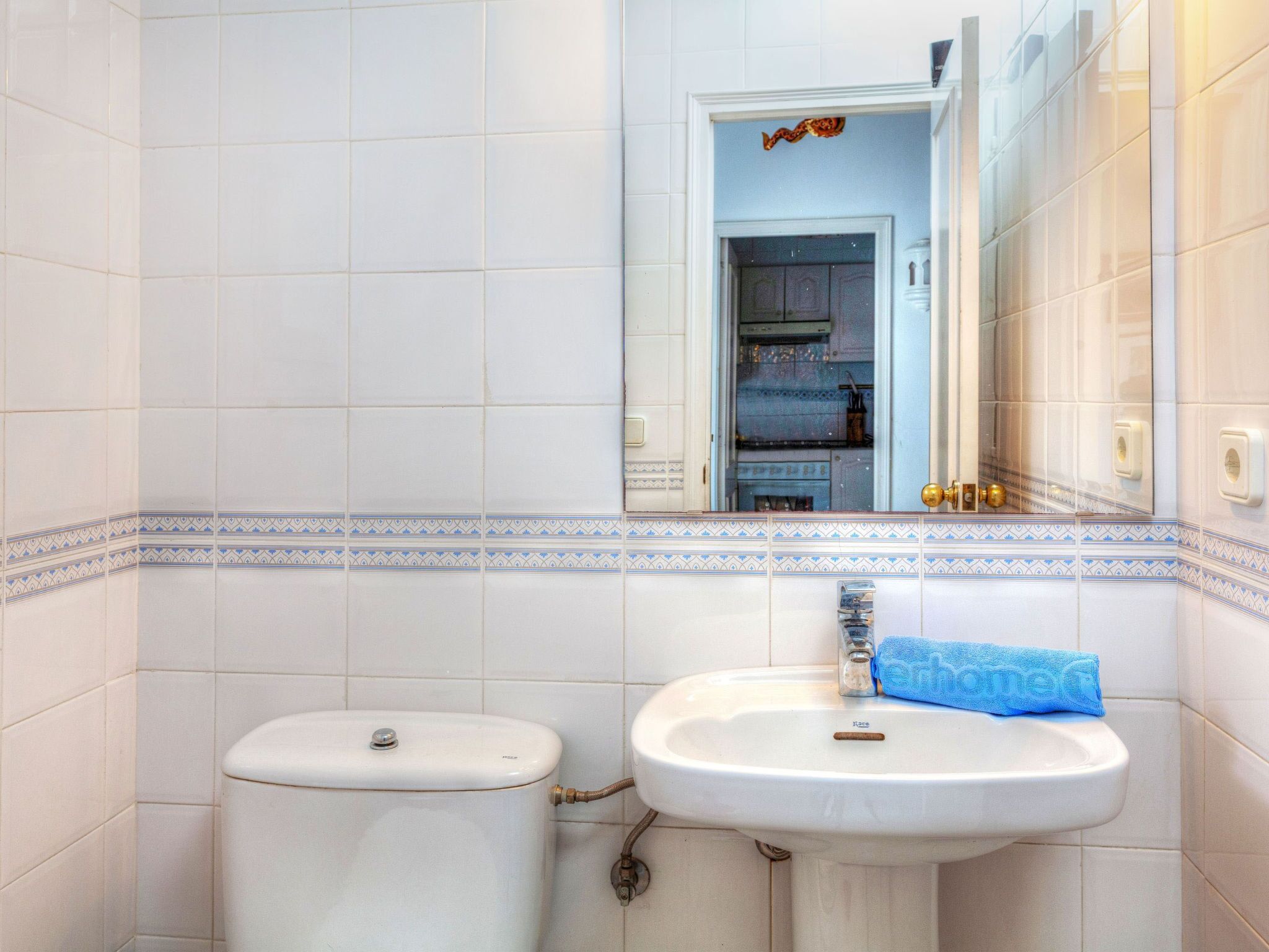 A bathroom with white tiles, a toilet, a sink with a towel, and a mirror reflecting a door and shelves.