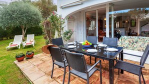 Un área de comedor al aire libre con una mesa dispuesta para cuatro personas, rodeada de sillas y vista al interior de la casa a través de puertas de vidrio.