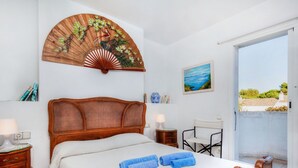 Un dormitorio con cama de madera, cabecero de ratán, ventilador de pared grande, mesita de noche con lámpara, una silla y vistas al exterior a través de una puerta corredera.