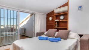 A hotel room with a bed, a wooden shelf with decorative items, a window with curtains, and a view of the city. - Port L'escala by Interhome (L'Escala)