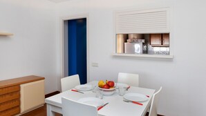 Un comedor con una mesa blanca para cuatro, una puerta azul y una cocina visible a través de una ventana.