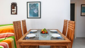 A dining area with a wooden table set for four, surrounded by wooden chairs. There are framed artworks on the walls, a small shelf with a mirror, and a colorful striped sofa. - Riells Park by Interhome (L'Escala)