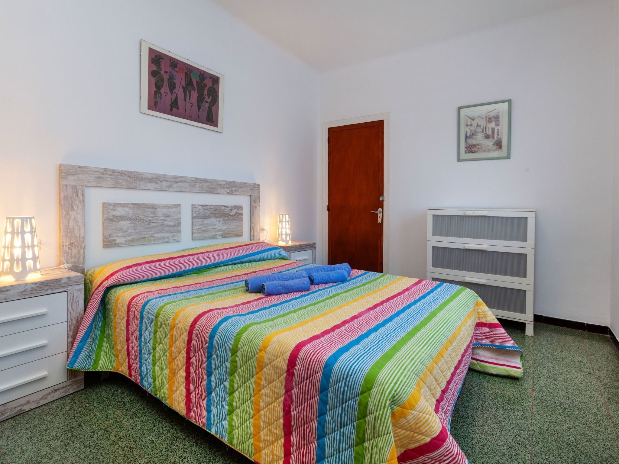 Ein Schlafzimmer mit einem farbenfrohen, gestreiften Bettüberwurf, einem hölzernen Kopfteil, einer weißen Kommode, einer Tür und einem gerahmten Bild an der Wand.