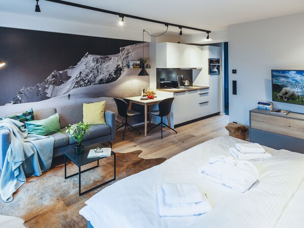 A modern hotel room with a large bed, a sofa, a desk, and a kitchen area.