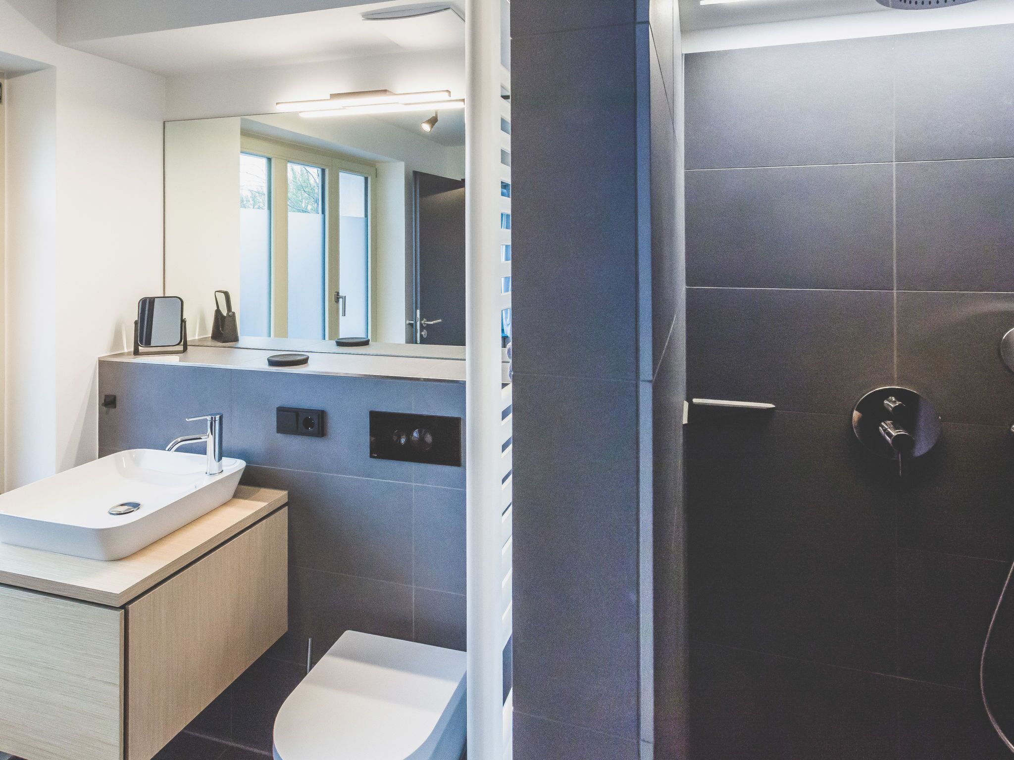 A modern bathroom with a white sink, a mirror, and a dark shower area.