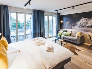 A modern hotel room with a large bed, a sofa, a coffee table, and a view of the outdoors.