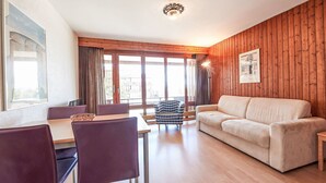 Uma sala de estar moderna com uma parede de madeira, um sofá bege, uma mesa de jantar com cadeiras e janelas amplas.