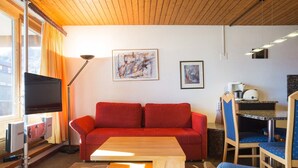A modern living room with a red sofa, a wooden coffee table, and a TV on a stand. There is a desk with a chair and a small kitchenette. - Dixence 212 by Interhome (Thyon-Les Collons)