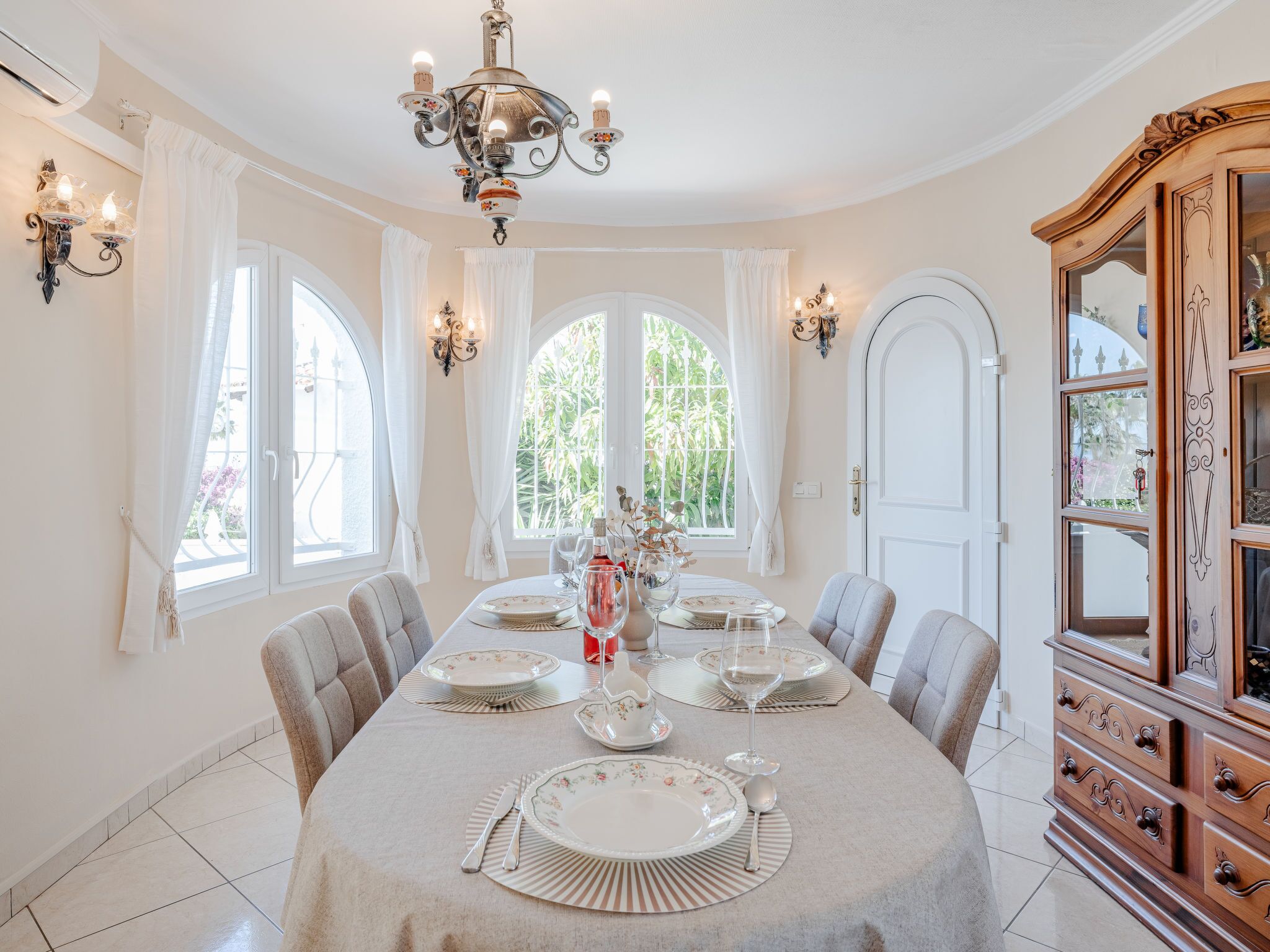 Une salle à manger avec une table ronde dressée pour six personnes, entourée de chaises. Un lustre est suspendu au-dessus de la table, et un buffet en bois avec des portes en verre se trouve à droite.