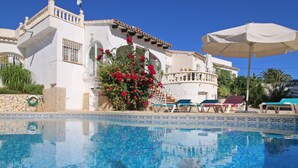 A clear blue pool with lounge chairs and umbrellas, surrounded by a white building with red flowers and a stone wall. - Heringklee by Interhome (Calpe/Calp)