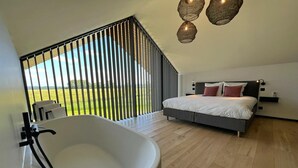 A modern bedroom with a large bathtub, a bed with pillows, and a view of a green field through a window with vertical blinds. - Romantic Stay with Views (Enneille)