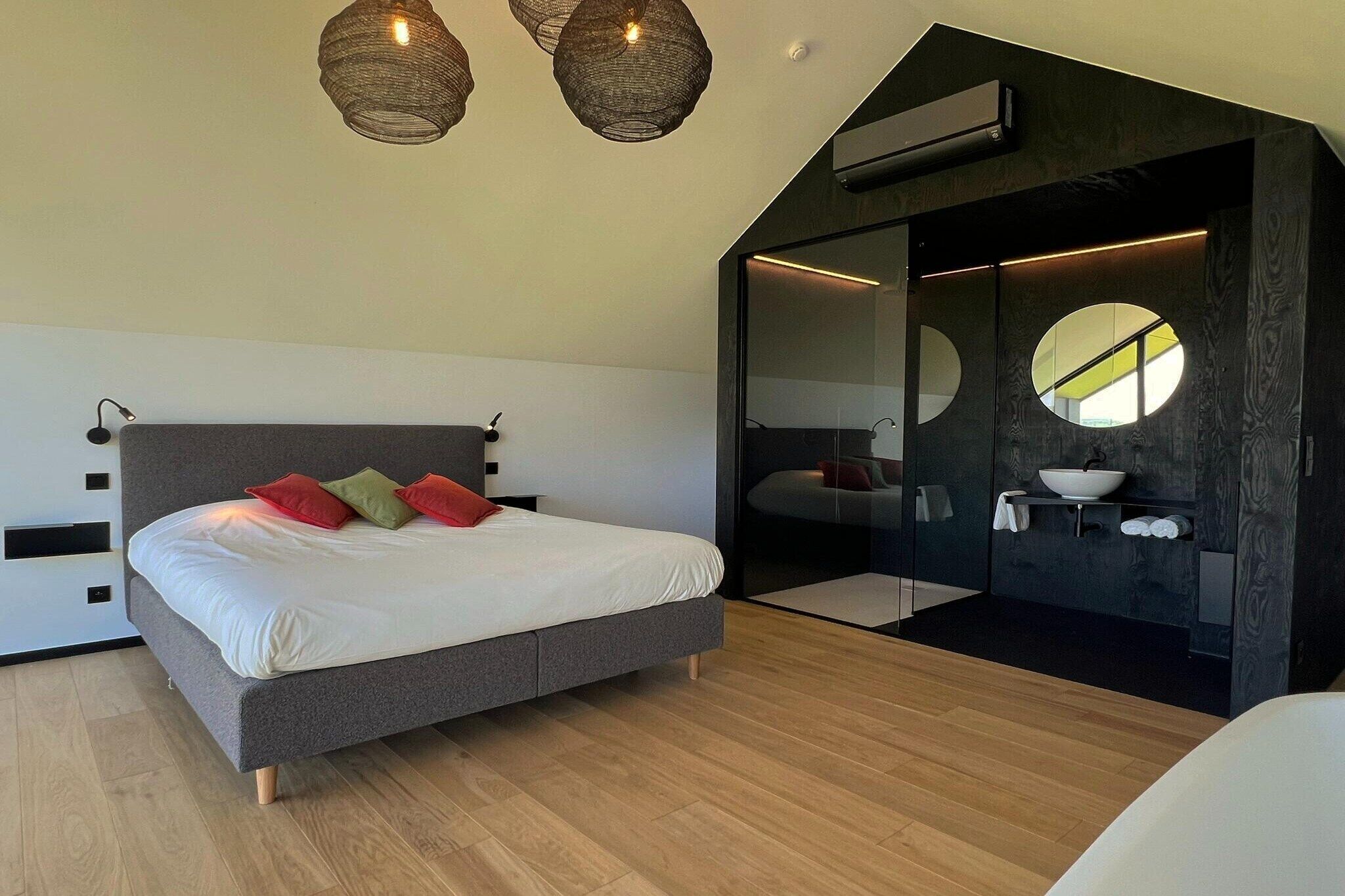 Una camera da letto moderna con un letto grande, un armadio a muro, uno specchio rotondo e un bagno con lavandino e porta asciugamani.