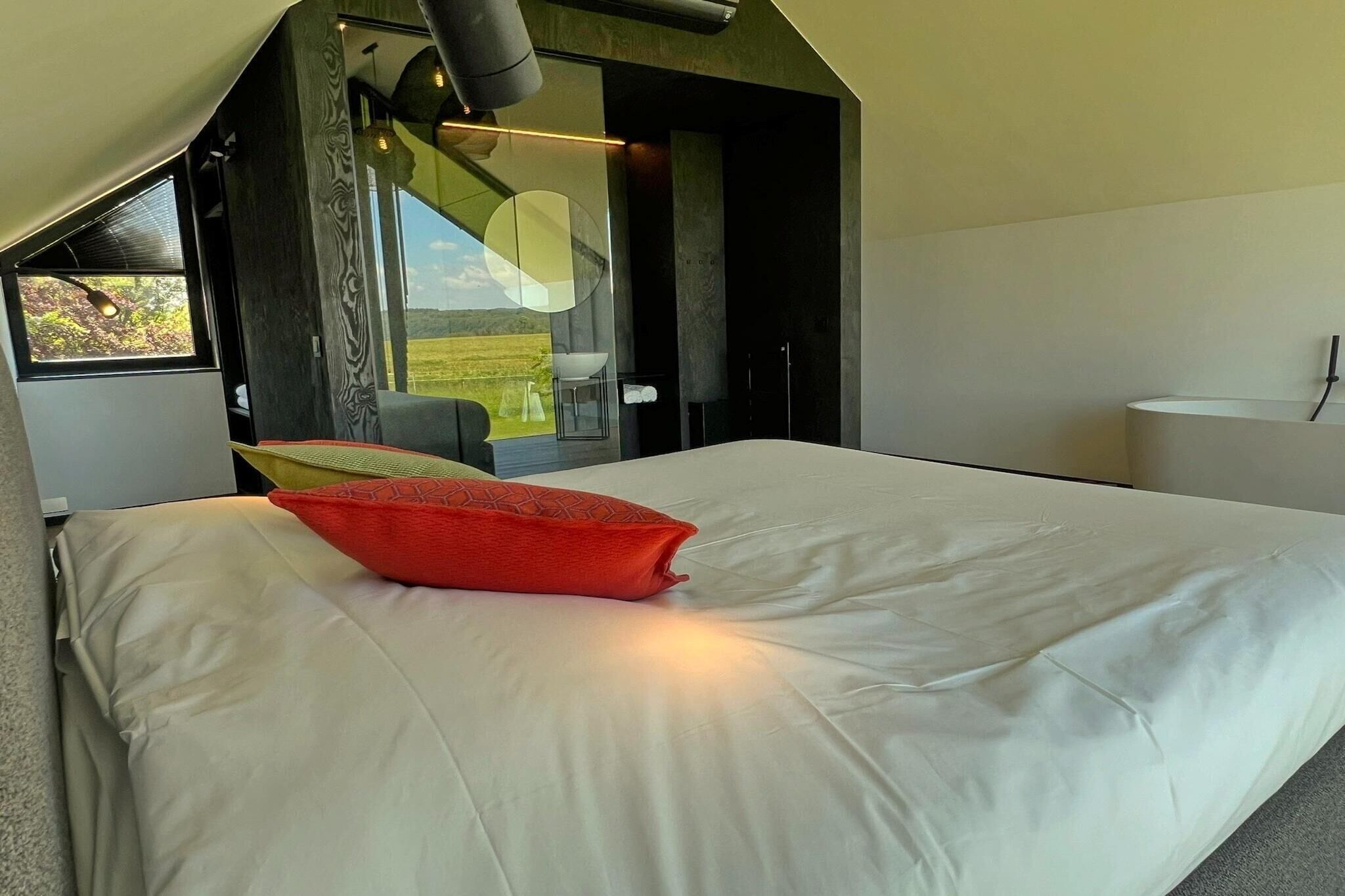 A modern bedroom with a large bed, a red pillow, and a view of a green landscape through a window.