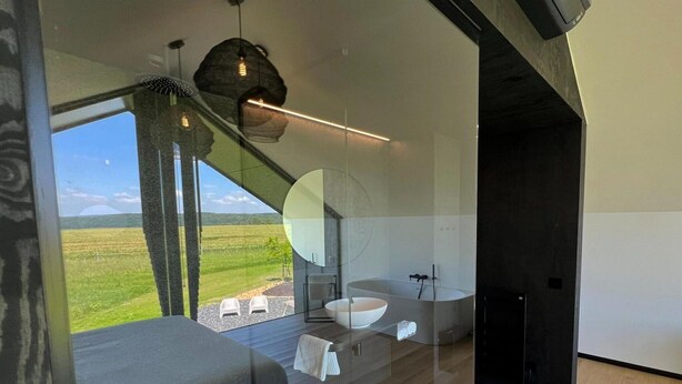 A modern bathroom with a large mirror, a white sink, and a bathtub. There is a view of a green landscape through large windows. - Cozy Romantic Stay Near Durbuy (Enneille)