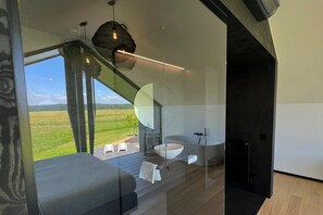 Une salle de bain moderne avec un grand miroir, un lavabo blanc et une baignoire. De grandes fenêtres offrent une vue sur un paysage verdoyant.