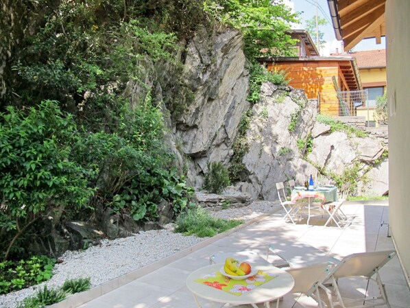 An outdoor patio with a table set for two, surrounded by greenery and a rocky backdrop.