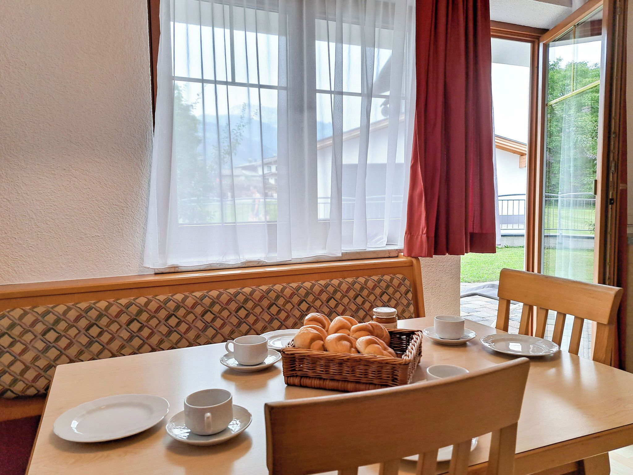 Uma área de refeições com uma mesa posta para dois, uma cesta de vime com pão e vista para um jardim pela janela.