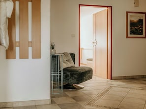 Una habitación con un sofá gris oscuro, una mesita de noche con una planta, una puerta que da a otra habitación y un cuadro en la pared.