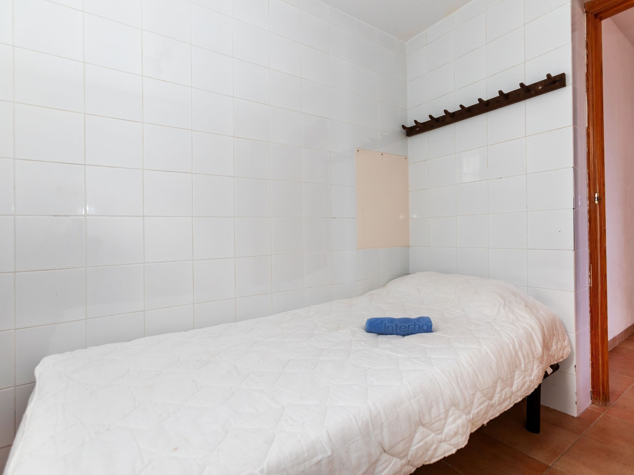 Une petite chambre avec un lit simple, des murs recouverts de carreaux blancs et un chambranle de porte en bois.