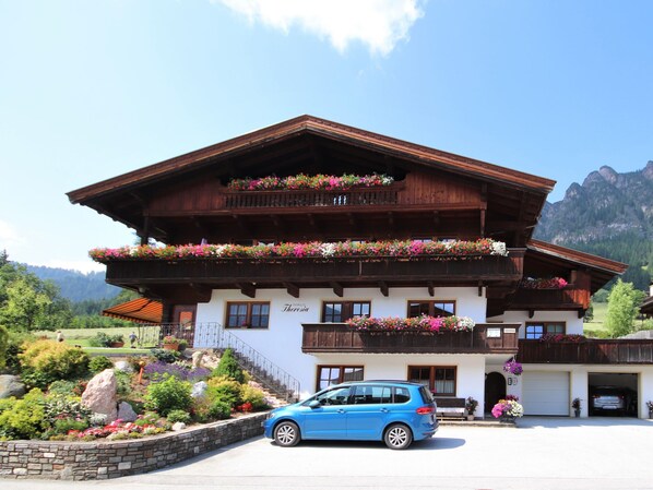 A traditional wooden chalet with balconies adorned with flowers, a car parked in front, and a mountainous backdrop.