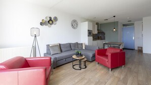 Een moderne woonkamer met een grijze bank, een rode fauteuil en een kleine ronde tafel met een plant. In de achtergrond is een keuken zichtbaar met een blauwe deur.