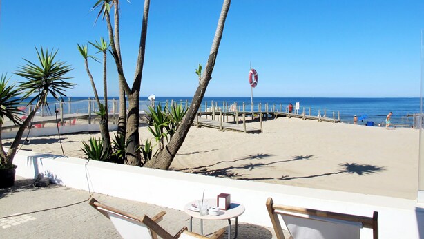 Un patio frente a la playa con sillas de madera y una pequeña mesa, con vista al océano.