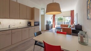 A modern kitchen with wooden cabinets, a dining table with red chairs, a hanging lamp, and a view of a balcony with a sofa and TV.