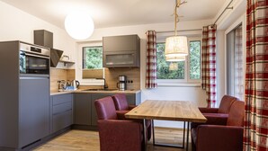 Una cocina compacta con electrodomésticos modernos, una zona de comedor con una mesa de madera y sillas rojas, y un ventanal con cortinas de cuadros.