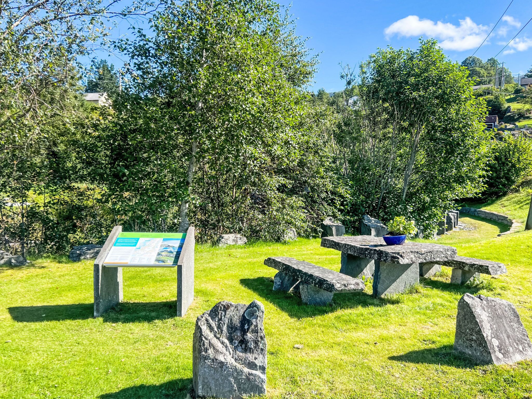 A picnic area with stone benches and a signboard, surrounded by lush greenery and trees.