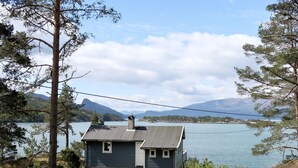 Une petite maison avec un toit en pente et une cheminée, située dans un espace verdoyant entouré d’arbres.