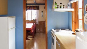 Une cuisine avec des armoires en bois, une cuisinière et un réfrigérateur. Il y a une fenêtre avec des rideaux et une étagère au-dessus de la cuisinière où sont rangées différentes bouteilles.