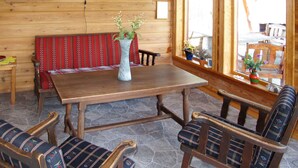 L’intérieur d’un chalet en bois est aménagé avec une table en bois, des bancs et des chaises. On y trouve des fenêtres avec des rideaux et des plantes en pot posées sur les rebords de fenêtre.