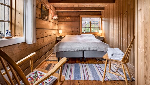 Ein gemütliches Schlafzimmer in einer Holzhütte mit einem großen Bett, zwei Nachttischen, einem Fenster mit Vorhängen, einem hölzernen Stuhl und einem bunten Teppich.