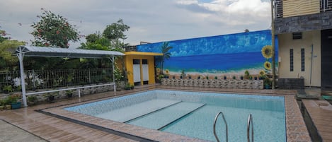A swimming pool with a tiled deck, a covered seating area, and a mural of a beach scene.