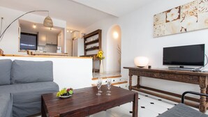 A modern living room with a sofa, a wooden coffee table with a fruit bowl, a flat-screen TV, and a dining area with a wooden table and chairs.