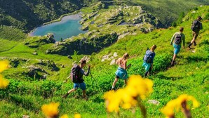 A group of hikers on a grassy mountain trail with a lake in the background.