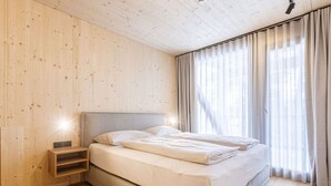 Un dormitorio con piso de madera, una cama con ropa de cama blanca, una mesita de noche y un ventanal con cortinas.