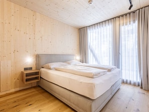Une chambre avec un plancher en bois, un lit avec une literie blanche, une table de chevet et une fenêtre avec des rideaux.
